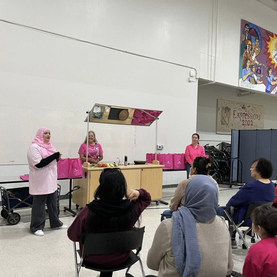 A woman speaks to an audience in a community center while two others stand behind a table with cooking supplies and pink bags. People are seated, listening. Colorful murals are on the wall.