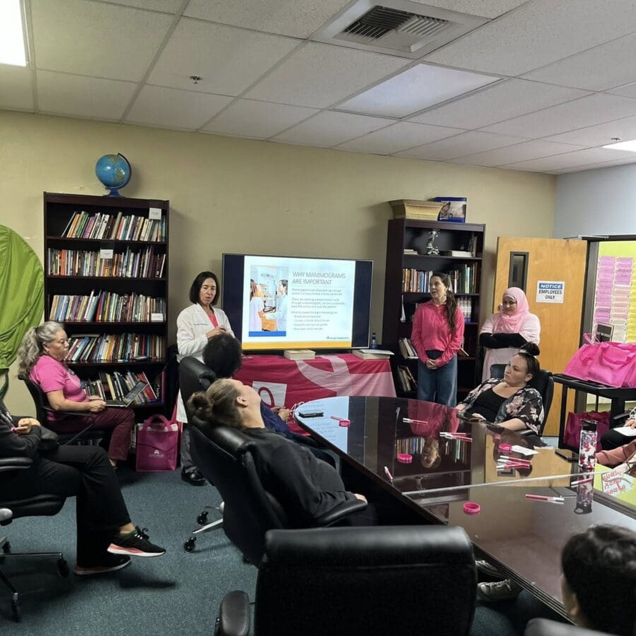 A cooking class with diverse participants seated in a community center. Instructors in pink shirts stand by a demonstration table with cooking items. Bright murals decorate the walls, creating a welcoming atmosphere.